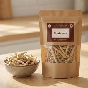 May include: A package of Shatavari, a pure Ayurvedic wellness product, next to a small bowl filled with the same dried herb. The package is brown with a clear window, and the bowl is ceramic. The text "Nature Spring" is visible.