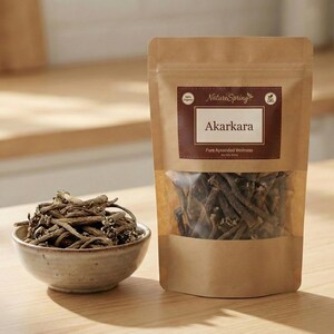May include: A brown paper bag of Akarakara, a pure Ayurvedic wellness product, with a clear window showing the contents. A small ceramic bowl holds a pile of the same root-like product. The label reads "Nature Spring".