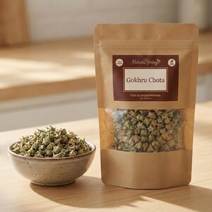 May include: A brown paper bag and a small ceramic bowl on a wooden surface. The bag has a clear window showing a quantity of green, spiky Gokru Chota. The bowl contains a similar quantity of the same product. The bag has a label that reads "Gokhru Chota".
