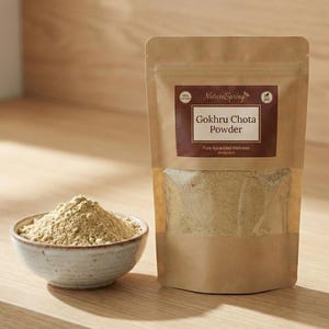 May include: A package of Gokhru Chota Powder and a small bowl of powder sit on a wooden surface. The package is brown with a label that reads "Gokhru Chota Powder" and "Pure Ayurved Wellness". The bowl is light-colored and filled with a beige powder.