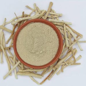 May include: Overhead shot of a small, round, terracotta-colored bowl filled with a fine, light beige powder. The bowl is surrounded by numerous pieces of dried, light-brown plant stems, all set against a white background.