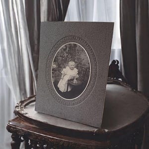 May include: A framed black and white photograph of a baby in a white dress, set in an oval frame. The frame is gray and stands on a dark wooden table with ornate carvings. The background includes curtains and a window.
