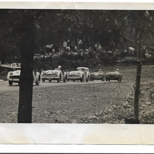 May include: A black and white photograph of a vintage car race. The photo shows several white cars driving on a winding road. The cars are all Triumph TR3s. The cars are driving past a crowd of spectators.