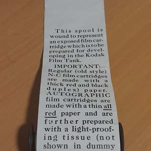 May include: A close-up of a vintage film spool with a label that reads "This spool is wound to represent an exposed film cartridge which is to be prepared for developing in the Kodak Film Tank. IMPORTANT: - Regular (old style) N-C film cartridges are made with thick red and black duplex) paper. AUTOGRAPHIC film cartridges are made with a thin all red paper and are further prepared with a light-proof-ing tissue (not shown in dummy spools) which permits of the record- "