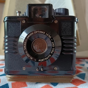 May include: A classic black camera featuring an "Envoy Optimax Lens". The camera has a rectangular shape with ribbed detailing and a viewfinder. The lens is inscribed with "Push In Views" and "Pull Out Portraits". A vintage photography item.