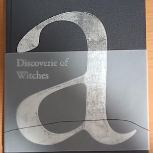 May include: A dark blue hardcover book with a silver foil design of the letter "a" on the front cover. The title "Discoverie of Witches" is printed in white on a white background.