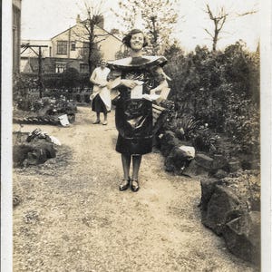 Könnte beinhalten: Vintage Schwarz-Weiß-Fotografie einer Frau in einem dunklen Kleid und dekorativem Kragen, die Papiere hält und auf einem Kiesweg steht. Eine andere Frau steht im Hintergrund. Die Szene ist ein Garten mit Bäumen und einem Haus.