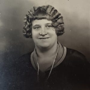 Könnte beinhalten: Schwarz-Weiß-Porträt einer Frau mit Bob-Frisur und Perlenkette. Die Frau lächelt und trägt ein dunkles Oberteil. Das Foto hat einen klassischen, zeitlosen Look.