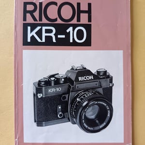 May include: A vintage camera manual for the Ricoh KR-10. The cover is a muted pink with the words "RICOH KR-10" in bold white letters. A black camera with a lens is featured on the front. The camera has the word "RICOH" on the top.