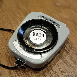 May include: A gray and black light meter with a silver dial and a black lens. The dial has markings for DIN, ISO, and f-stops. The lens has a white label that reads "AUTO TK-62".