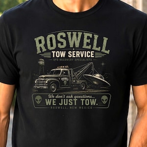May include: Black t-shirt featuring a graphic of a tow truck towing a UFO. The design includes the text "ROSWELL TOW SERVICE UFO RECOVERY SPECIALISTS" and "WE DON'T ASK QUESTIONS... WE JUST TOW."