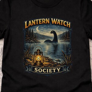 May include: Black t-shirt with a scenic design. The image shows a lake, mountains, and a creature. The text "Lantern Watch Society" is displayed. A vintage lantern is in the foreground.