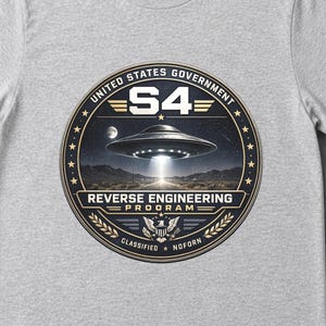 May include: A light gray t-shirt featuring a circular graphic with the text UNITED STATES GOVERNMENT S4 REVERSE ENGINEERING PROGRAM CLASSIFIED NOFORN. The design depicts a metallic flying saucer hovering over a desert landscape at night.