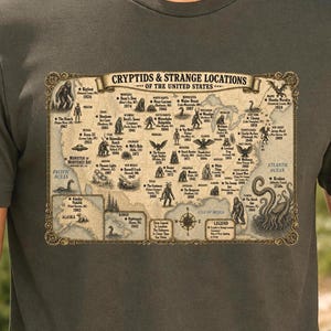 May include: A dark olive green t-shirt featuring a vintage-style map titled Cryptids & Strange Locations of the United States. The graphic displays various mythical creatures and mysterious events across the country with dates and locations.