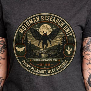 May include: Dark gray t-shirt featuring a circular graphic with the text "Mothman Research Unit" and "Point Pleasant, West Virginia." The graphic depicts a winged creature, a bridge, and a forest scene. The text "Cryptid Observation Team" and "Est. 1966" are also included.