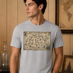 May include: A heather grey short-sleeve t-shirt featuring a vintage-style map graphic titled Cryptids & Strange Locations of the United States. The print displays various mythical creatures and geographic markers across a beige map of the country.
