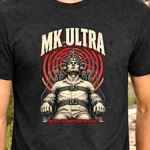 May include: A charcoal gray short-sleeve t-shirt featuring a graphic print of a person in a chair with a head apparatus. The text MK ULTRA appears at the top in white, and the text CONTROL THE MIND CONTROL REALITY appears at the bottom in red.