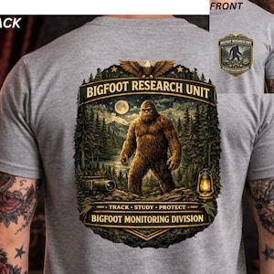 May include: A gray t-shirt featuring a Bigfoot graphic on the back and a smaller version on the front. The back graphic includes a Bigfoot illustration, text that reads "BIGFOOT RESEARCH UNIT" and "BIGFOOT MONITORING DIVISION".