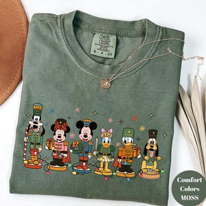 May include: A moss green short-sleeve t-shirt with a crew neckline and a graphic print of six Disney characters dressed as nutcrackers. The characters include Goofy, Minnie Mouse, Mickey Mouse, Daisy Duck, Donald Duck, and Pluto with string lights.