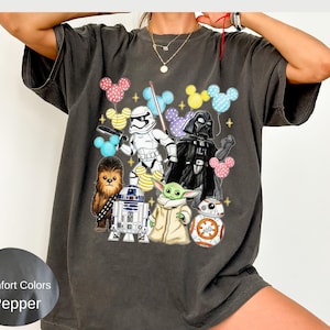 May include: A charcoal gray short-sleeve t-shirt featuring a graphic print of Star Wars characters including Darth Vader, a Stormtrooper, Chewbacca, R2-D2, BB-8, and Grogu, surrounded by colorful patterned Mickey Mouse ear balloons and gold stars.