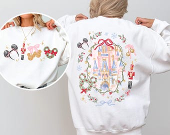 May include: A white crewneck sweatshirt featuring a watercolor-style print of a pink castle surrounded by a green wreath, a red nutcracker, and holiday icons. The front chest area displays smaller matching graphics of ears, bells, and a wreath.