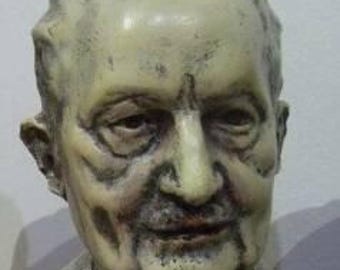 May include: A bust sculpture of a man with a neutral expression, wearing a suit and tie. The sculpture is primarily beige and gray, with visible details in the face and clothing. The bust is mounted on a dark rectangular base.