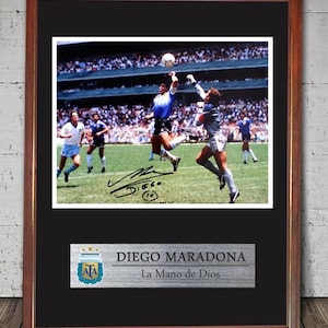 May include: Framed photograph of a soccer game featuring Diego Maradona. The image shows Maradona jumping to hit the ball with his hand. The frame is brown with a black mat. A plaque below reads "DIEGO MARADONA La Mano de Dios".