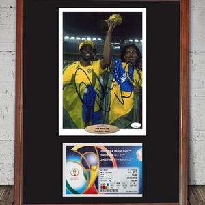 May include: Framed print featuring a photograph of two soccer players in yellow jerseys, one holding the World Cup trophy. Below is a ticket from the 2002 FIFA World Cup. The frame is brown with a black mat.
