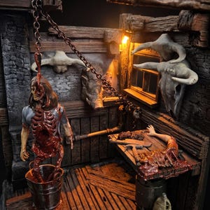 May include: A detailed diorama depicting a gruesome scene. A figure with exposed internal organs hangs from a chain. A table displays severed limbs and meat. The setting is a rustic, wooden structure with animal skulls.