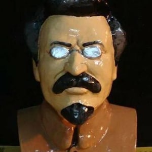 May include: A painted bust of a man with a stern expression. The sculpture features a tan face, black hair, a large black mustache, and a goatee. The figure wears glasses and a brown collared shirt.