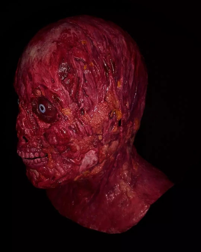 Pu&ograve; includere: Una maschera realistica e raccapricciante che raffigura un volto in decomposizione con carne esposta e un singolo occhio visibile. La maschera &egrave; principalmente rossa con aree gialle e bianche, su uno sfondo nero. La bocca &egrave; aperta, rivelando i denti.