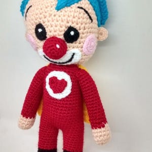 Pu&ograve; includere: Bambola pagliaccio all'uncinetto fatta a mano. La bambola ha un corpo rosso, scarpe nere e capelli blu. Il pagliaccio ha un naso rosso, guance rosa e un cuore bianco sul petto. La bambola ha un mantello giallo.