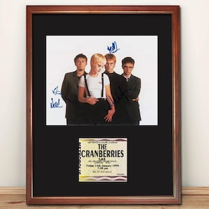 May include: Framed photograph featuring The Cranberries band members, with signatures. Beneath the photo is a concert ticket from Metropolis Music, LAS, dated 14th January 1994, with the text "The Cranberries". The frame is brown.