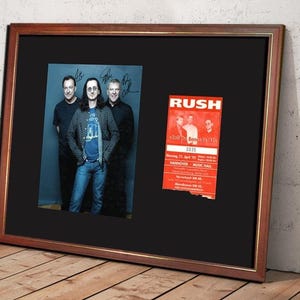 Könnte beinhalten: Gerahmtes Foto der Band Rush mit Unterschriften und einem Konzert-Ticket-Ausschnitt. Das Foto zeigt drei Männer vor einem blauen Hintergrund. Das Konzertticket ist rot mit weißem Text. Der Rahmen ist braun.