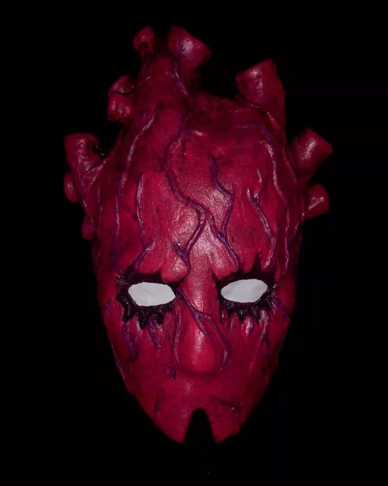 Peut inclure: Un masque fait &agrave; la main, de couleur cramoisie, avec un design en forme de c&oelig;ur. Le masque pr&eacute;sente des orbites blanches, des d&eacute;tails violets en forme de veines et une surface textur&eacute;e. Le masque est pos&eacute; sur un fond noir.