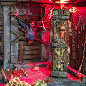 May include: A detailed diorama of a horror scene with a red-lit interior. The scene includes a wooden door adorned with skulls and chains, a skeleton, and a figure seemingly trapped. Skulls and candles are scattered throughout.