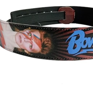 May include: A black guitar strap featuring a portrait of David Bowie with red and blue accents. The word "BOWIE" is printed in blue. The strap has a black backing and a brown leather end piece.