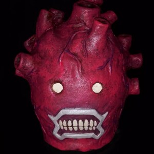 Peut inclure: Un masque rouge en forme de c&oelig;ur avec un design mena&ccedil;ant. Le masque pr&eacute;sente des yeux blancs, une bouche bord&eacute;e d'argent avec des dents blanches et des veines saillantes. Le masque est pos&eacute; sur un fond noir.