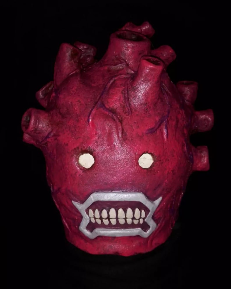 Peut inclure: Un masque rouge en forme de c&oelig;ur avec un design mena&ccedil;ant. Le masque pr&eacute;sente des yeux blancs, une bouche bord&eacute;e d'argent avec des dents blanches et des veines saillantes. Le masque est pos&eacute; sur un fond noir.
