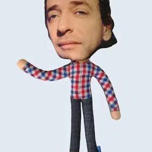 May include: A fabric doll with a large head featuring a man's face. The doll wears a red, white, and blue plaid shirt and dark denim pants. The doll's arms are outstretched, and it has a small blue tag attached to one leg.
