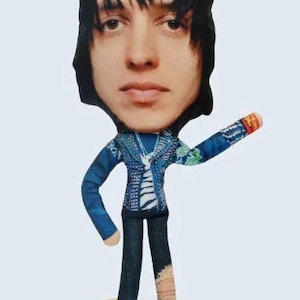 May include: A fabric doll with a printed face and black hair. The doll wears a blue denim jacket, dark blue jeans with a ripped knee, and a yellow tag that says "CIRCO". The doll's arm is outstretched.