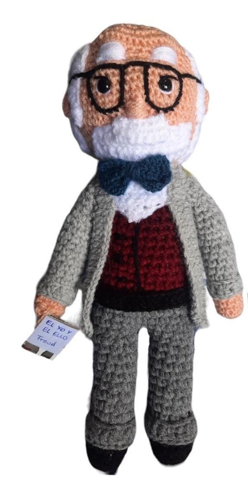May include: Crocheted doll of a man with glasses, a white beard, and a bow tie. He wears a grey jacket over a burgundy vest and grey trousers. A small tag reads "El Yo y El Ello Freud."