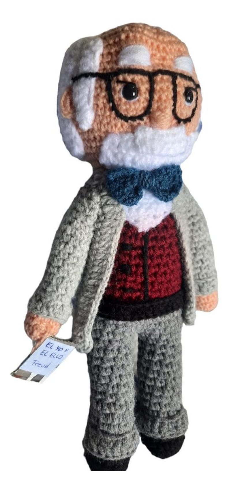 May include: Crocheted doll of a man with glasses, a white beard, and a blue bow tie. He wears a grey suit jacket over a red waistcoat and grey trousers. A small tag reads "El Yo y El Ello Freud."