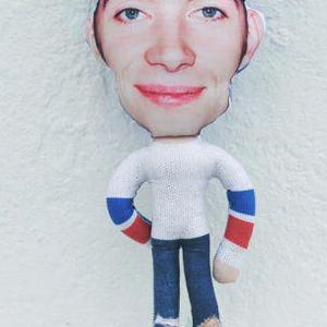May include: A handmade doll with a printed face and a white sweater with blue and red striped sleeves. The doll wears blue jeans with distressed details. A pink tag is attached to the doll's leg.