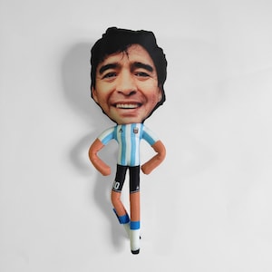 May include: A plush doll of a smiling man with dark hair, wearing a light blue and white striped soccer jersey, black shorts, and white socks. The doll has brown arms and legs, and the number 10 is visible on the shorts.
