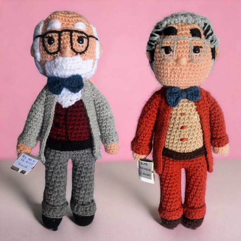 May include: Two crocheted dolls of older men, one in a grey suit and the other in a red suit. Both dolls wear spectacles and bow ties, and hold small cards with text. The background is a solid pink.