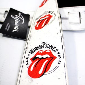 May include: White guitar strap with the Rolling Stones' red lips logo and the text "FIFTY YEARS". The strap has a black backing and a tag that says "Liguana".