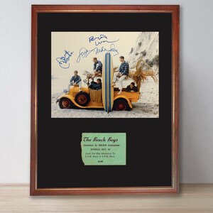 May include: Framed vintage photograph of The Beach Boys with surfboards on a yellow truck, signed by the band. A green ticket stub below the photo reads "The Beach Boys" and event details.