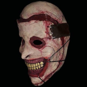 Puede incluir: Una máscara grotesca de color blanquecino con una banda de cuero cosida en la frente. La máscara presenta una sonrisa amplia e inquietante con dientes amarillos, cuencas de ojos delineadas en rojo y detalles de sangre simulada, creando un tema de terror.