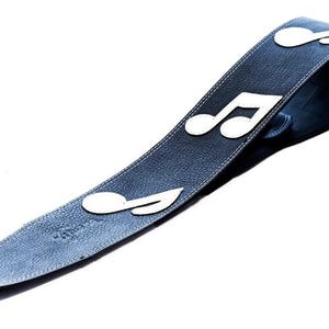May include: A dark blue guitar strap with white musical note cutouts. The strap is made of leather and has white stitching along the edges. The musical notes are evenly spaced along the strap.